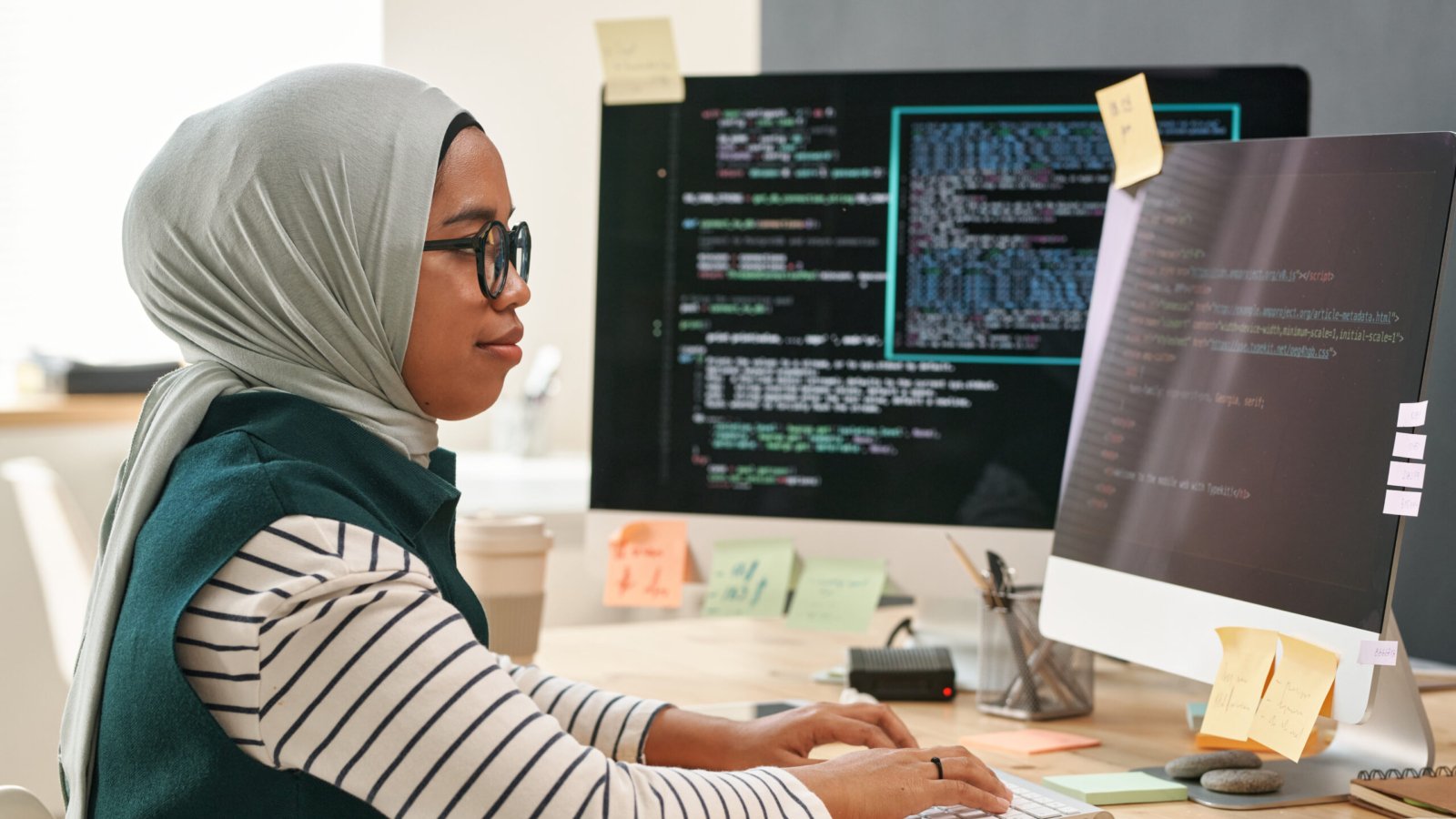 Young Muslim businesswoman in hijab looking at computer screen while typing and decoding data in front of monitor in office