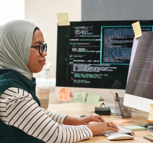 Young Muslim businesswoman in hijab looking at computer screen while typing and decoding data in front of monitor in office